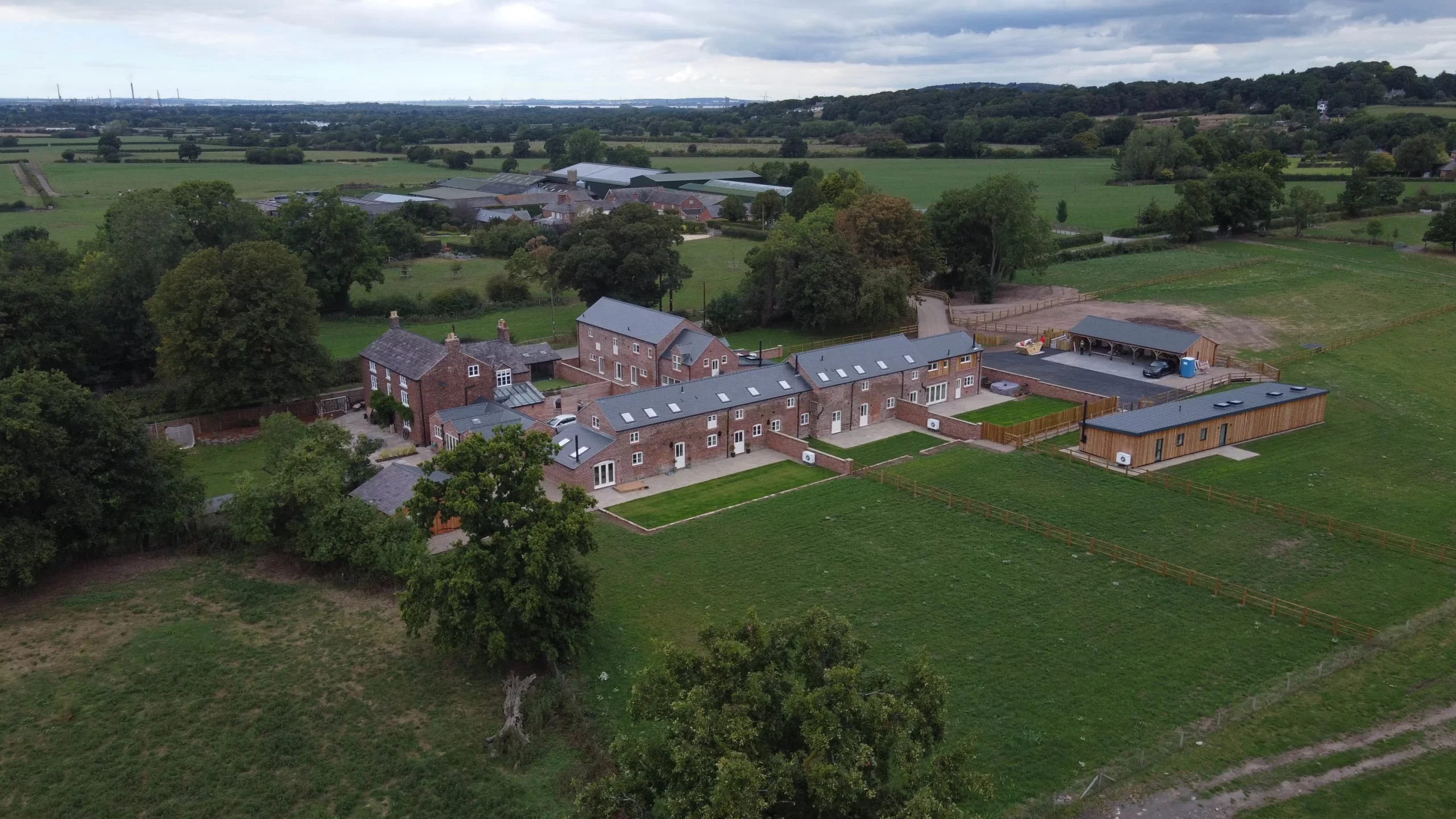 Aerial view of a countryside farm complex with brick cottages, long converted barns, outbuildings and fenced green fields surrounded by trees.