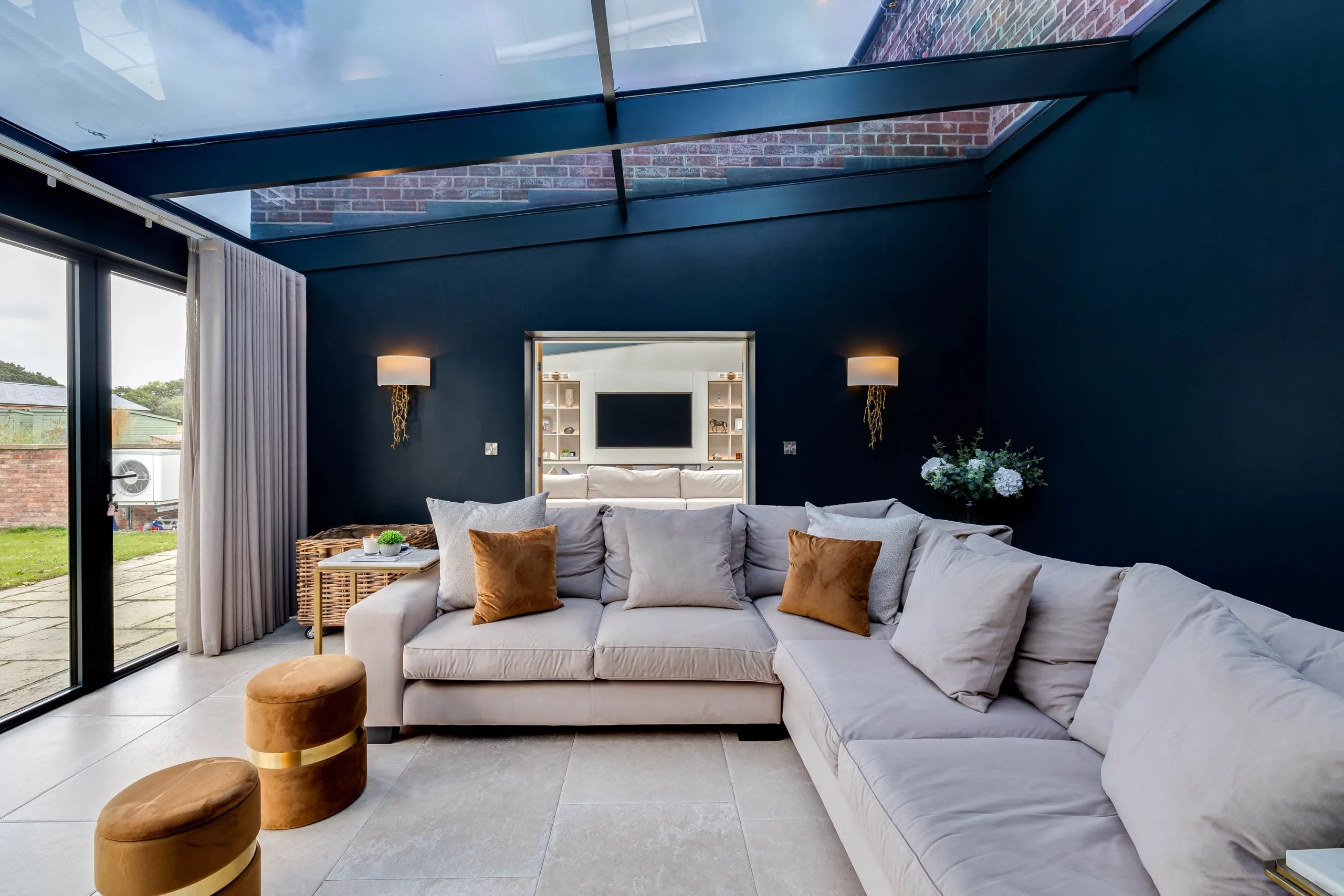 Spacious modern living room with a large pale grey sectional sofa, brown velvet cushions, glass skylight and dark navy feature wall.