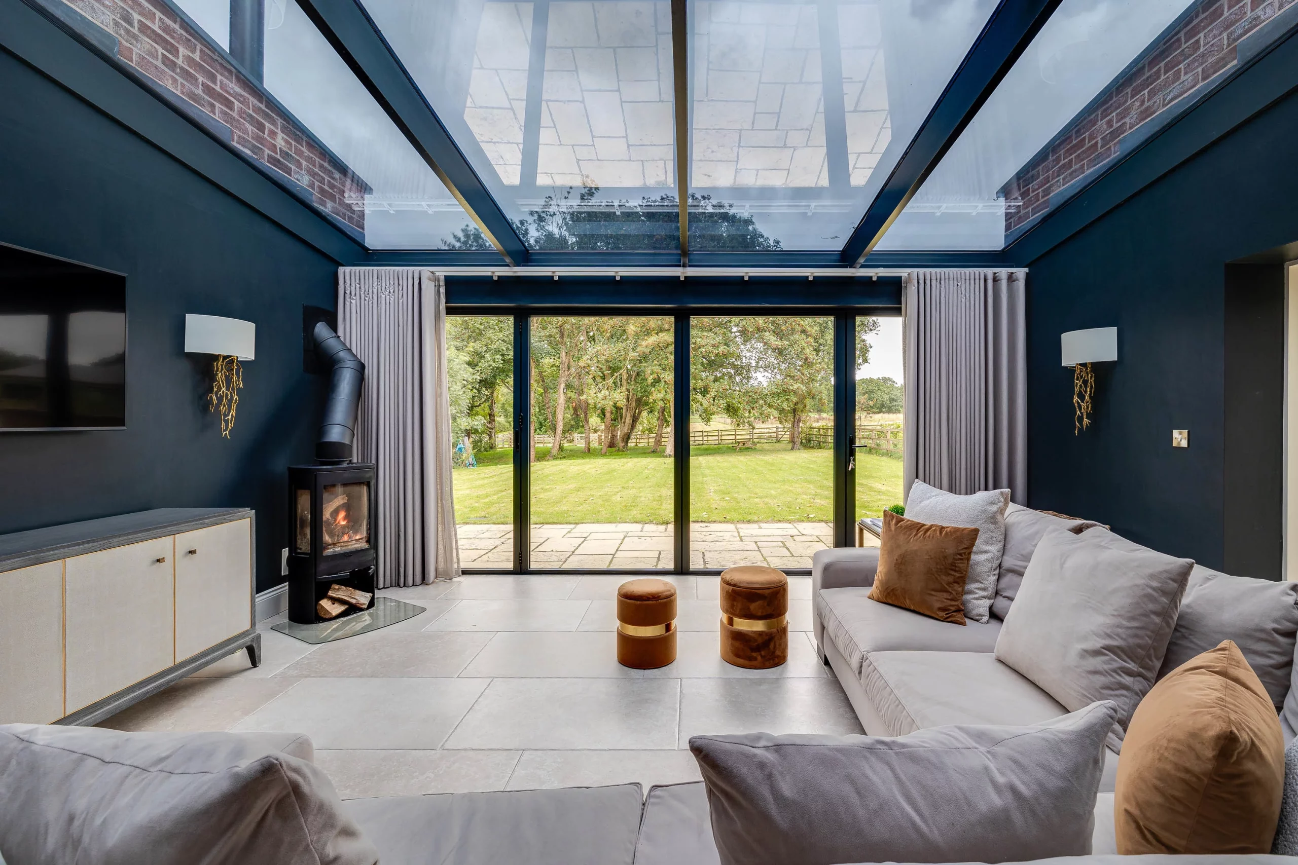 Contemporary living room with glass roof, sliding doors opening to a green lawn, grey sofas and a wood-burning stove