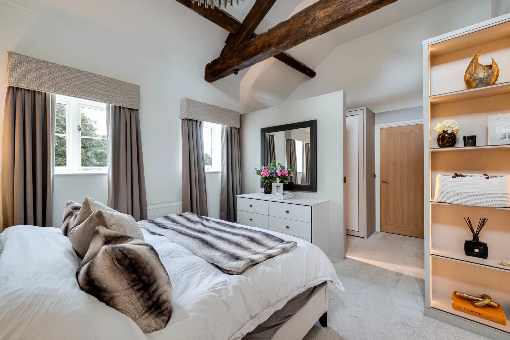 Bright bedroom with exposed wooden beams, neutral bed linens and faux-fur throws, dresser with mirror and built-in shelving.