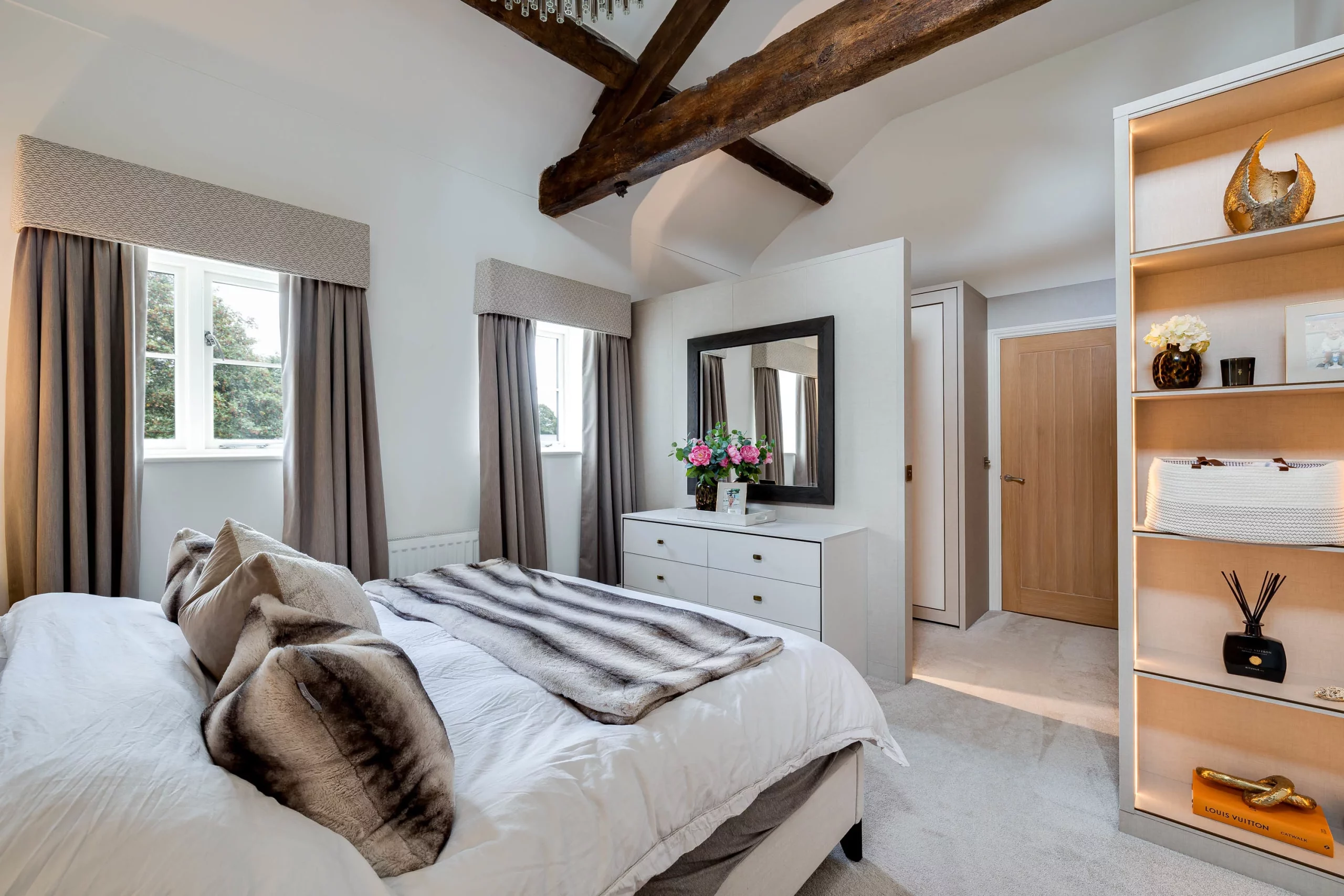 Bright bedroom with exposed wooden beams, neutral bed linens and faux-fur throws, dresser with mirror and built-in shelving.