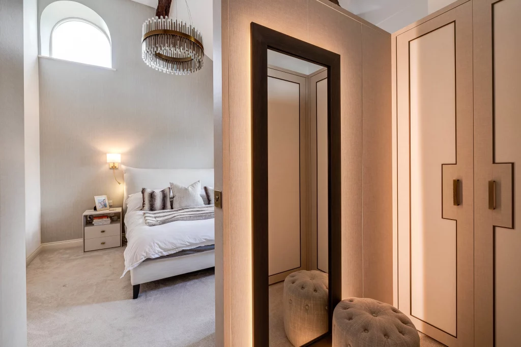Neutral-toned bedroom seen through doorway with tufted bench, full-length mirror, built-in wardrobe and chandelier above bed