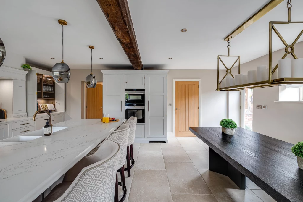 Modern kitchen with marble island, upholstered bar stools, wooden beam ceiling, pendant lights and a dark dining table with potted plants