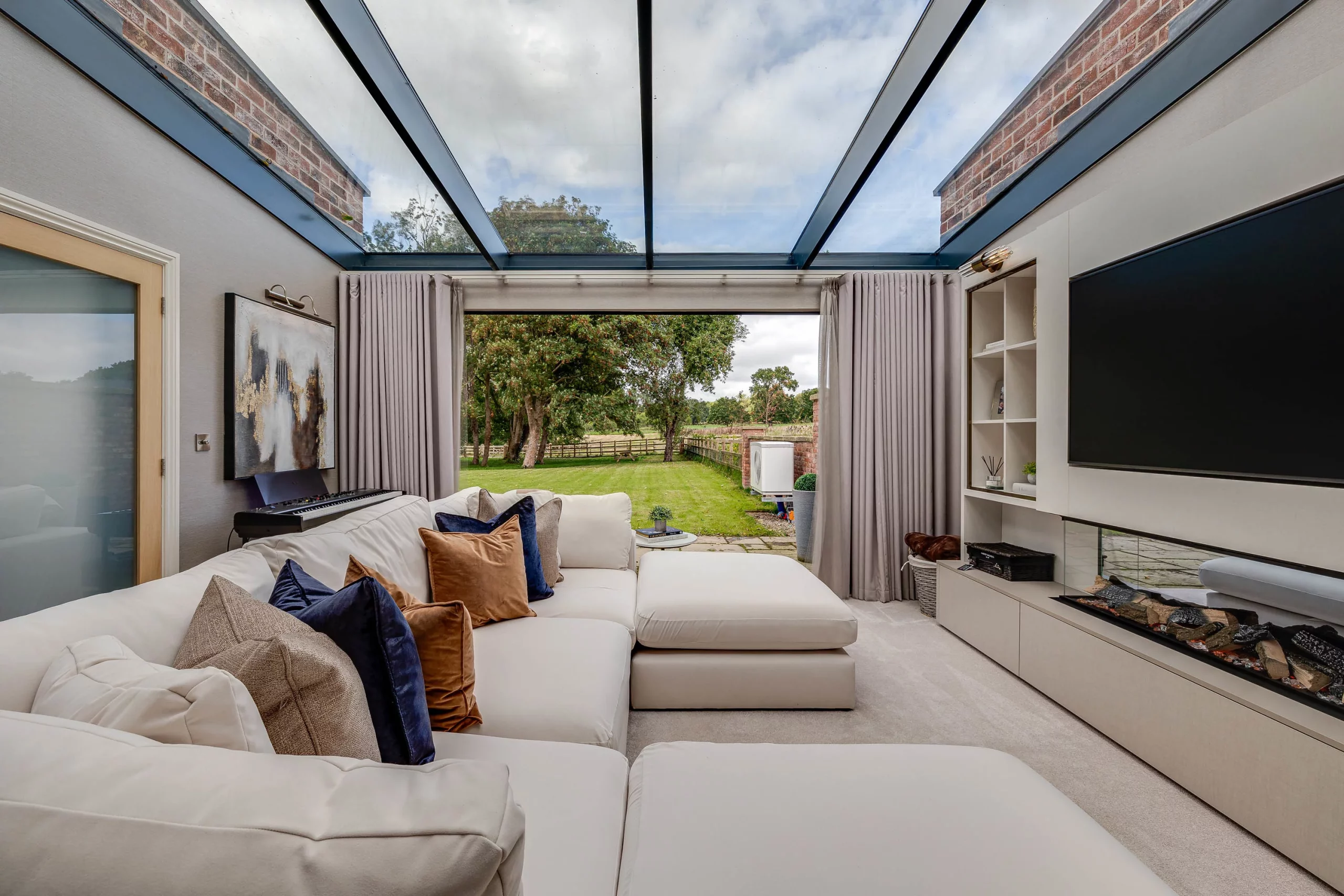 Spacious modern living room with cream sectional, blue and brown cushions, glass roof and open view to a green backyard and trees