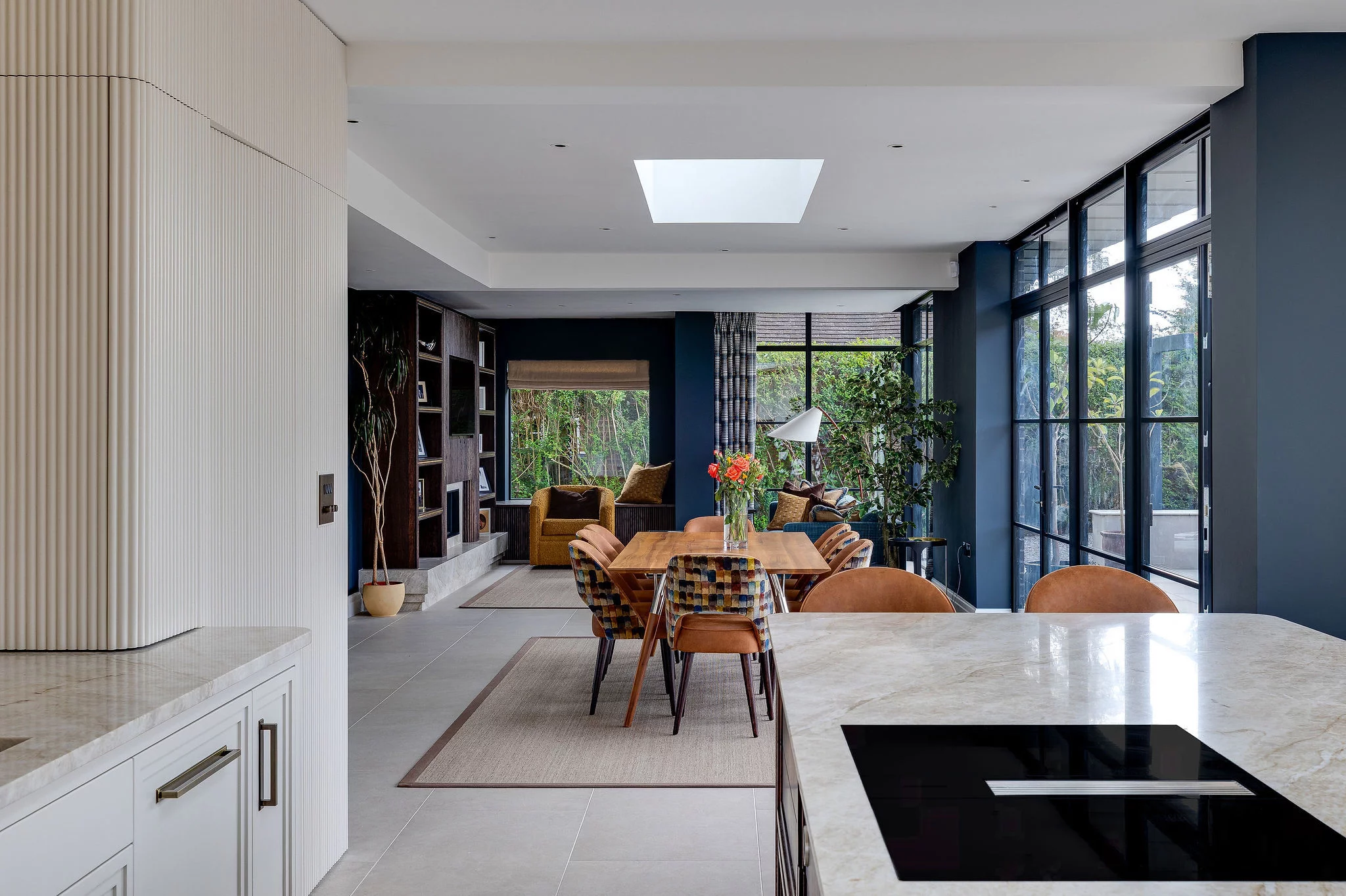 Open-plan dining and living area with a long wooden table, mixed-pattern chairs, blue walls and floor-to-ceiling windows.