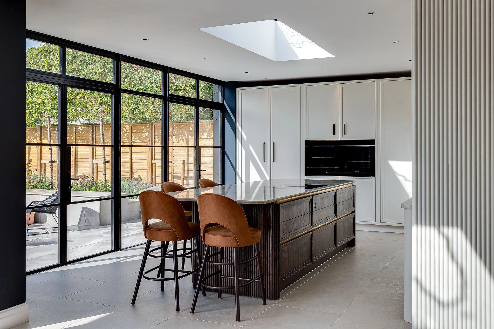 Modern kitchen with a dark wood island, four brown leather bar stools, floor-to-ceiling black-framed windows and white cabinetry
