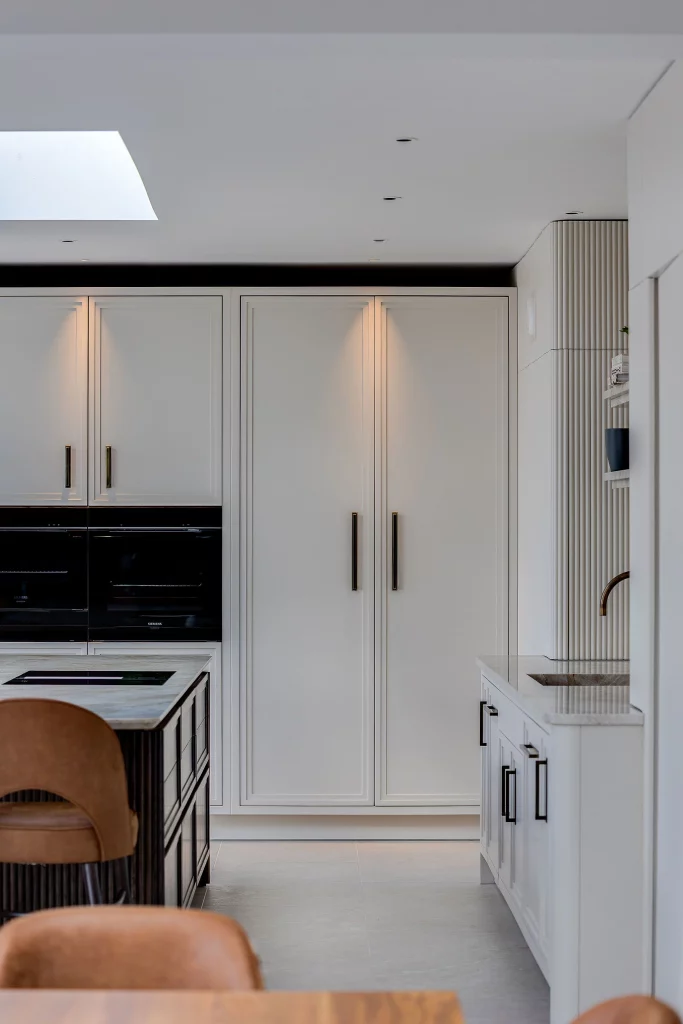Tall white kitchen cabinets with black handles, built-in ovens, marble island edge and tan leather bar stools in modern kitchen