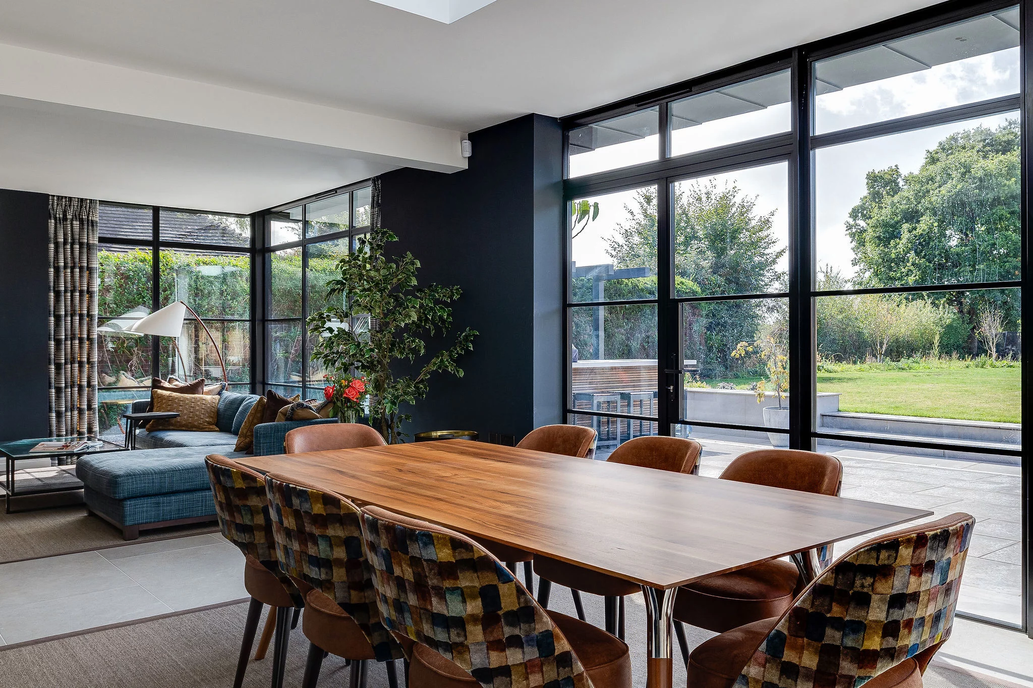 Sunlit modern dining and lounge area with a long wooden table, patterned upholstered chairs, blue sectional sofa, large floor-to-ceiling windows and garden view