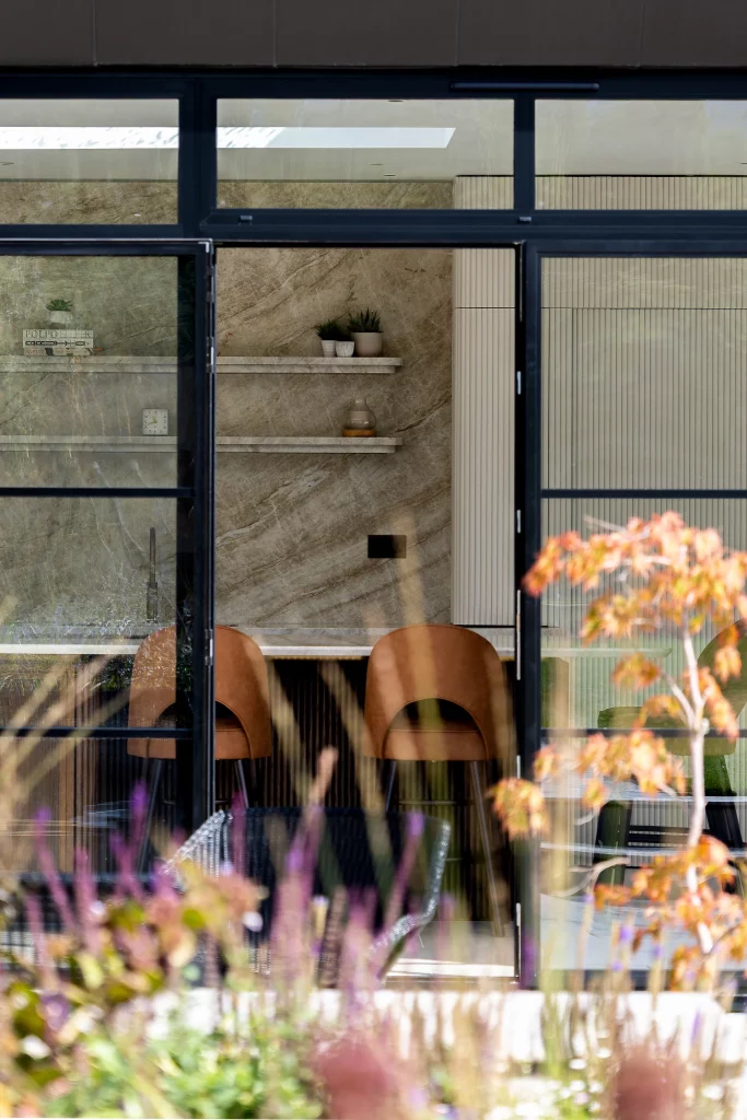 Window view into a modern dining area with two brown leather chairs, marble shelves, potted plants and blurred garden in foreground