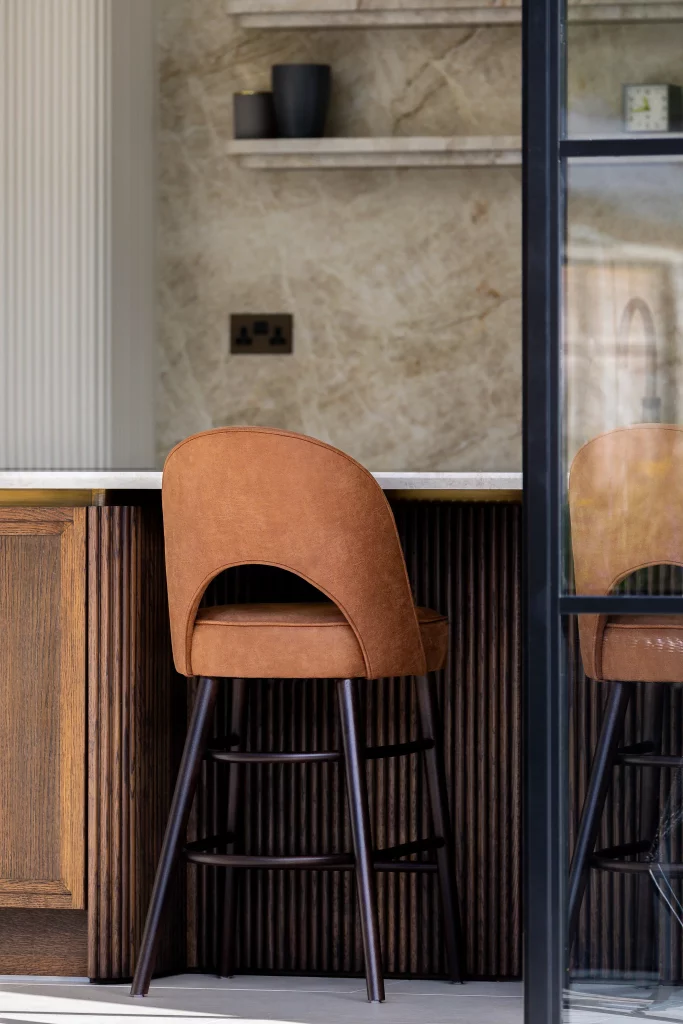 Tan leather bar stool with curved back and dark wooden legs tucked under a marble-topped wooden counter, reflected in glass.