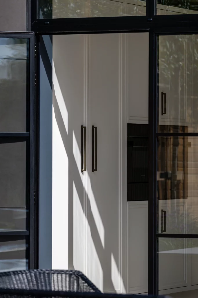 Narrow view of white pantry doors with long black bar handles seen through black-framed glass window, shadow patterns on wall