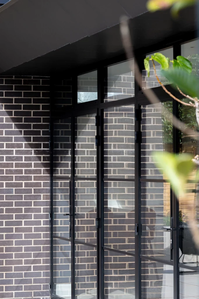 Tall black-framed glass doors and windows set into a dark grey brick wall with leafy plant blurred in foreground