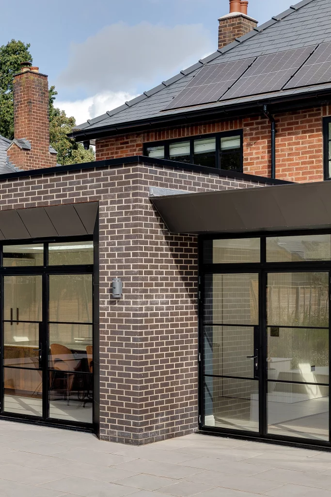 Corner of a modern brick house with large black-framed glass doors, flat-roof extension and solar panels on the slate roof