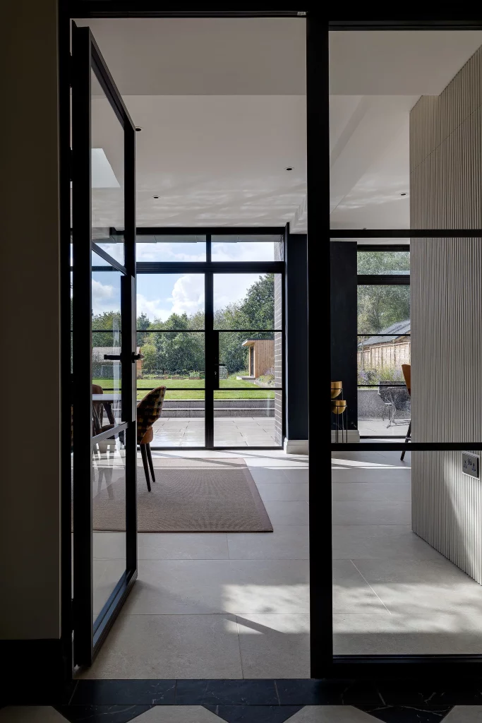 Open black-framed glass doors leading into a bright modern room with large windows showing a grassy garden and pavilion beyond.