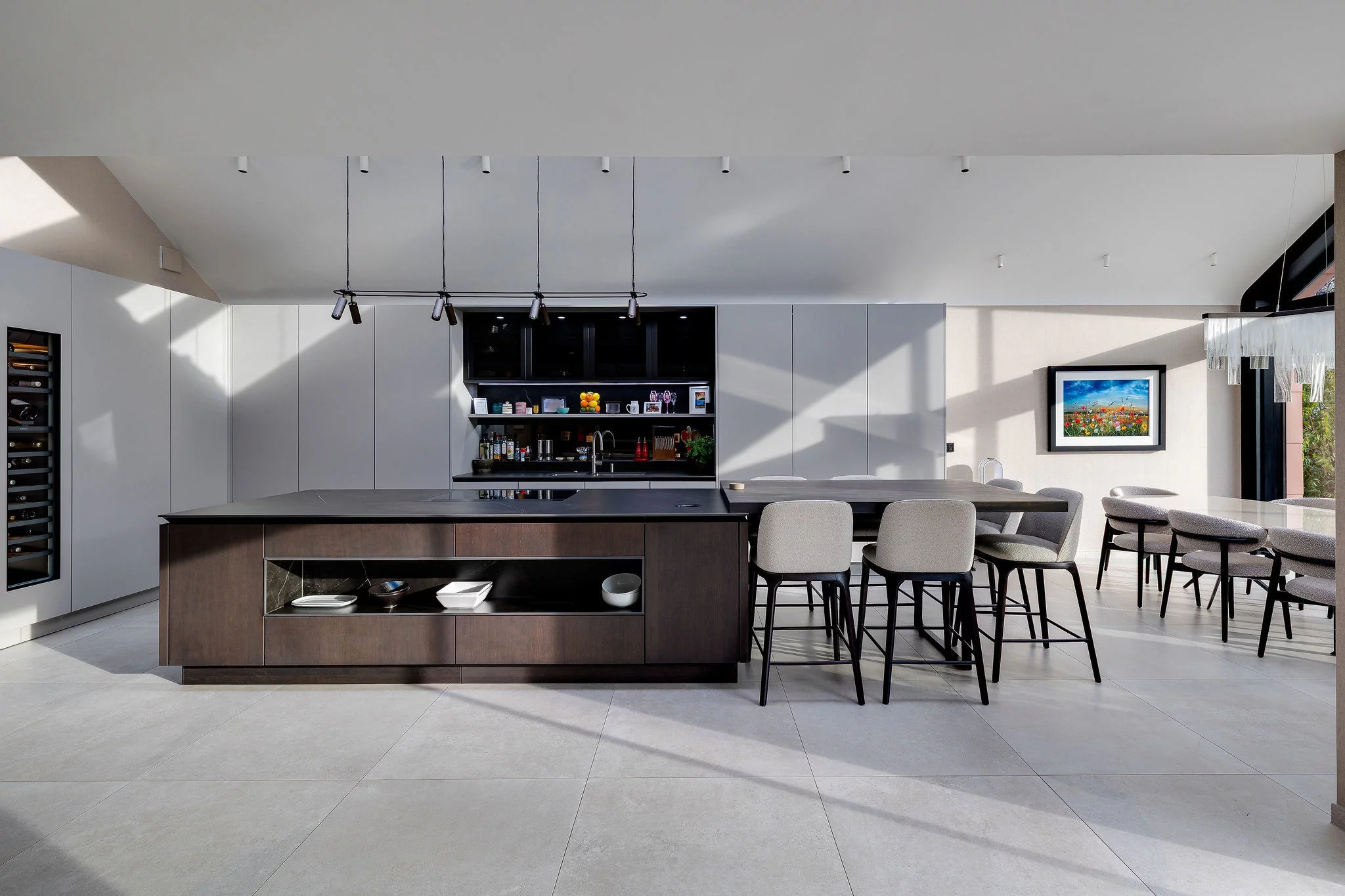 Modern open-plan kitchen with a large dark island, integrated shelving, four grey bar stools, pale tile floor and a dining table to the right.