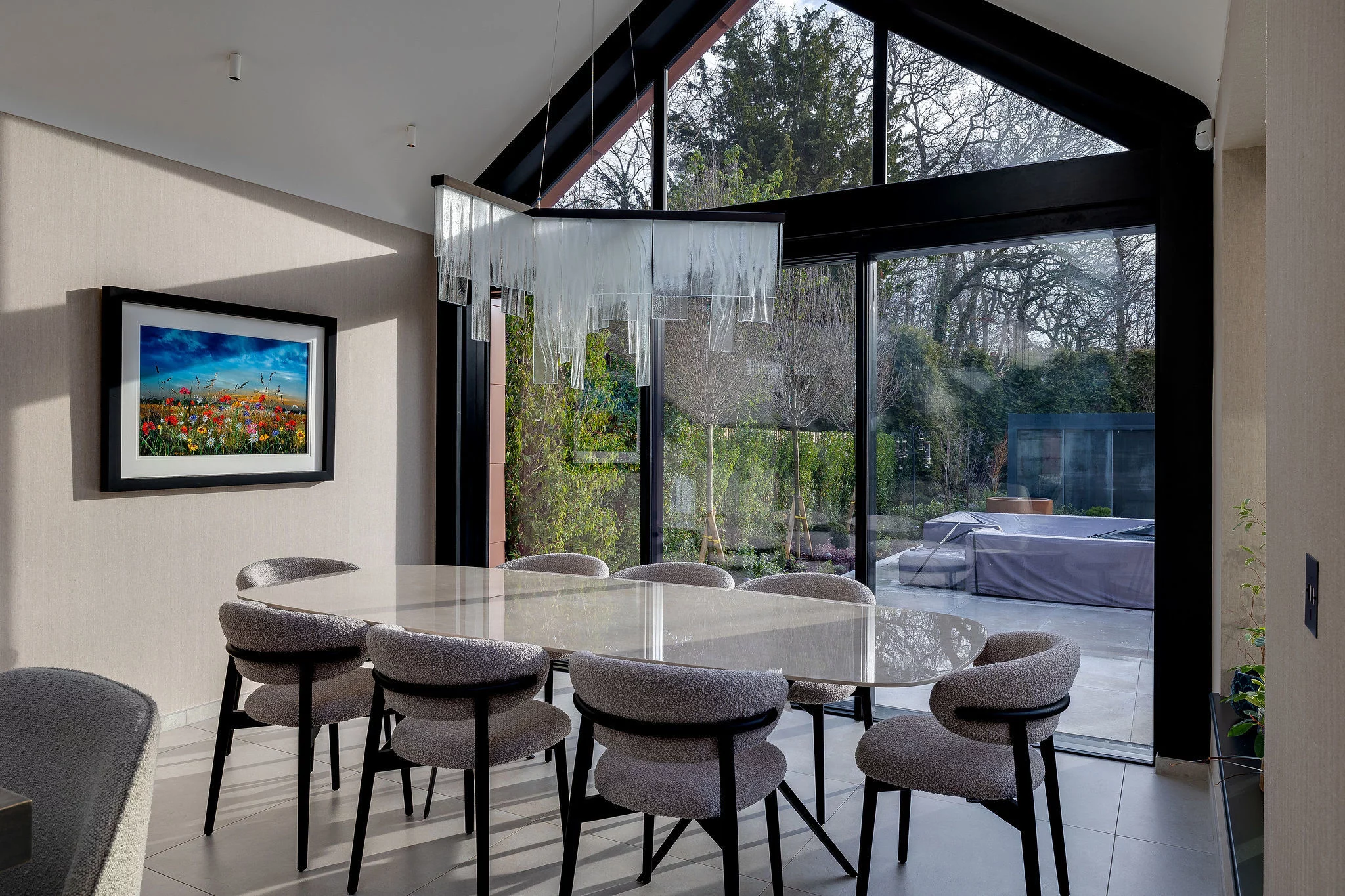 Contemporary dining room with oval marble table, eight upholstered gray chairs, glass pendant chandelier and large floor-to-ceiling windows overlooking a garden.