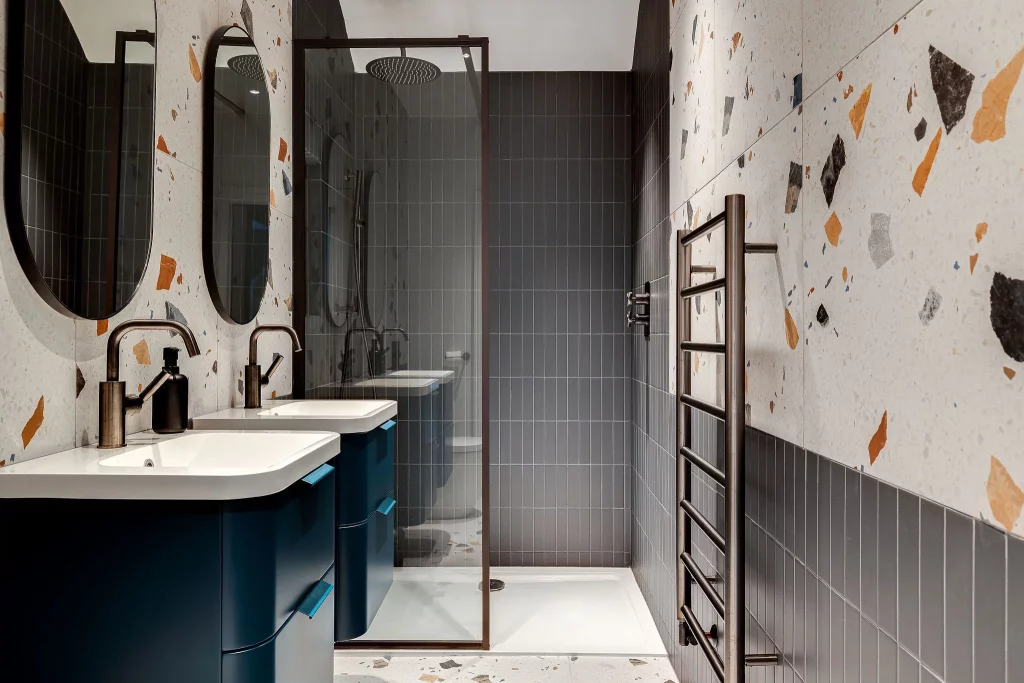Modern bathroom with twin sinks and round mirrors, a glass-enclosed shower with rain head, terrazzo-patterned walls and a heated towel rail