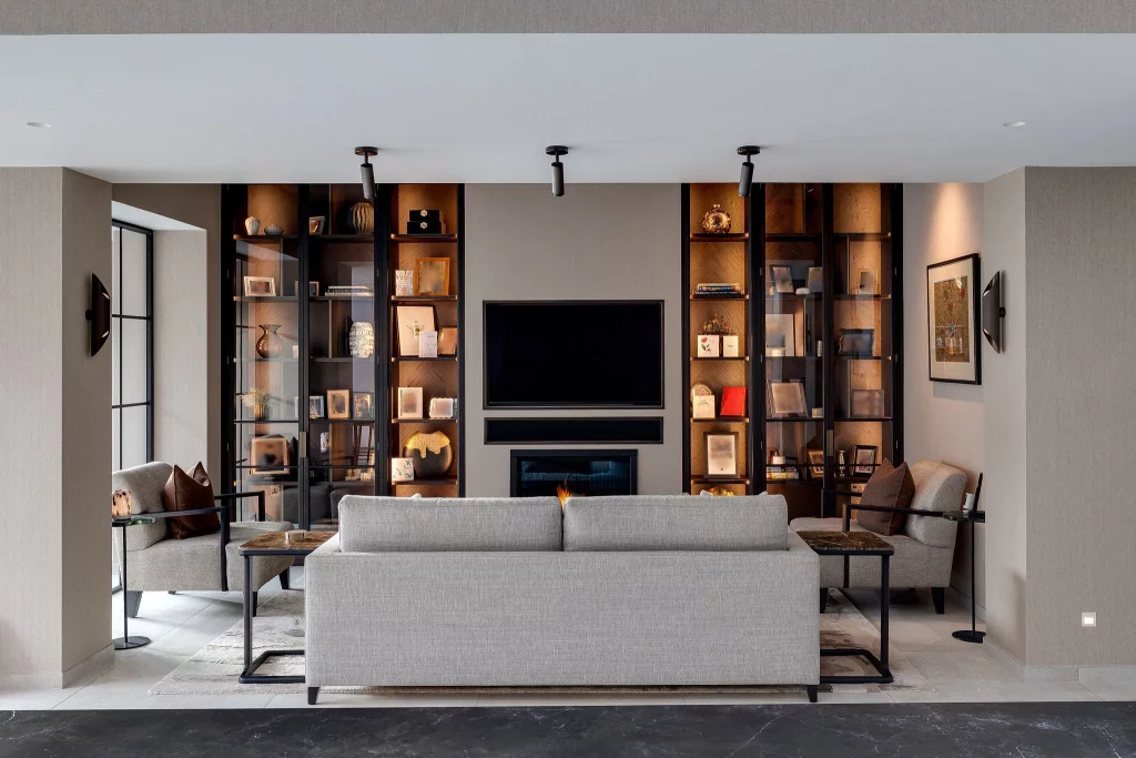 Symmetrical modern living room with grey sofa facing a wall-mounted TV and flanked by lit glass display shelves and armchairs