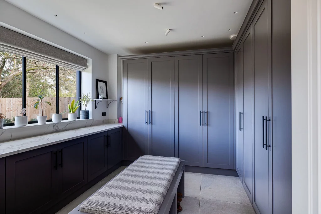 Walk-in dressing room with grey fitted wardrobes, a cushioned bench, marble-effect countertop and potted plants on the window sill.