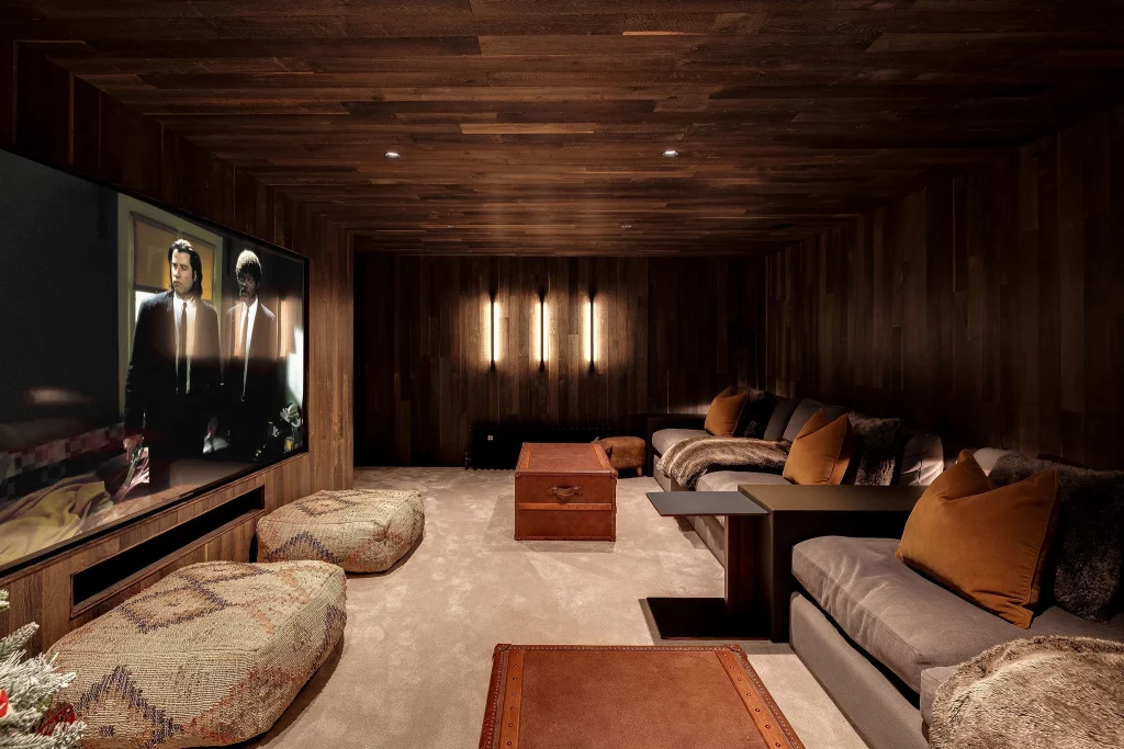 Cozy home cinema with dark wood walls, large screen, low poufs, leather trunk coffee table and plush grey sofas with brown cushions
