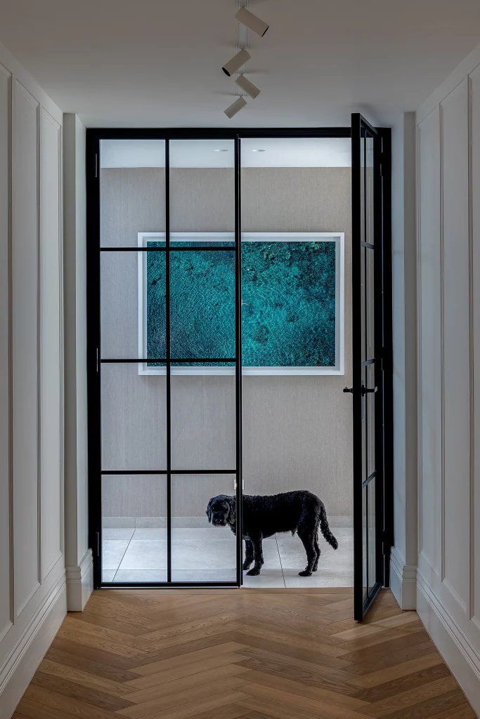 Black dog standing behind a tall black-framed glass door in a modern hallway with herringbone wood floor and teal artwork.