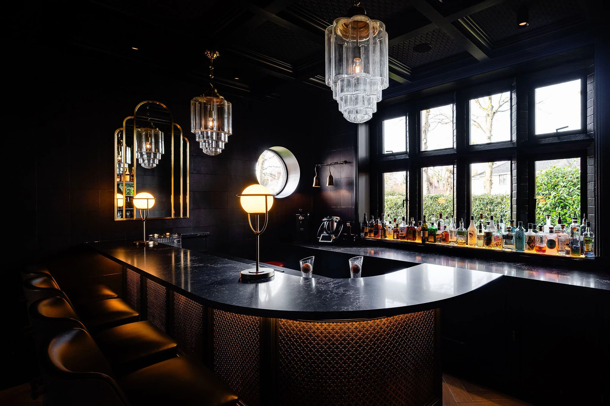 Dimly lit home bar with curved black marble counter, glowing globe lamps, crystal pendant lights, leather bar stools and bottles lined on a window sill