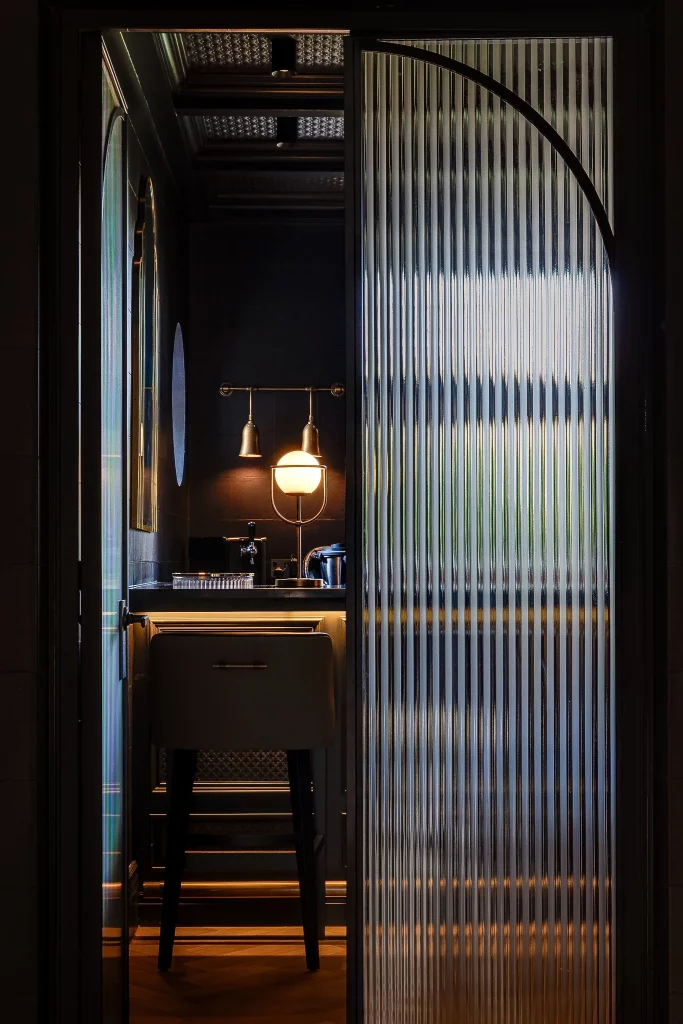 Narrow dimly lit vanity area seen through a ribbed glass sliding door, with a lit globe lamp, mirror and stool.