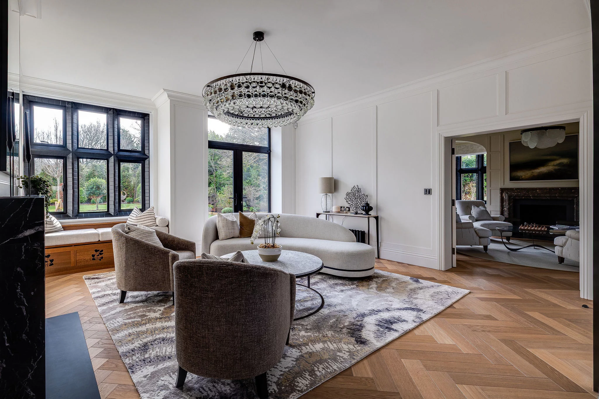 Bright modern living room with curved cream sofa, two brown armchairs around a round coffee table on a patterned rug, large black-framed windows and crystal chandelier.