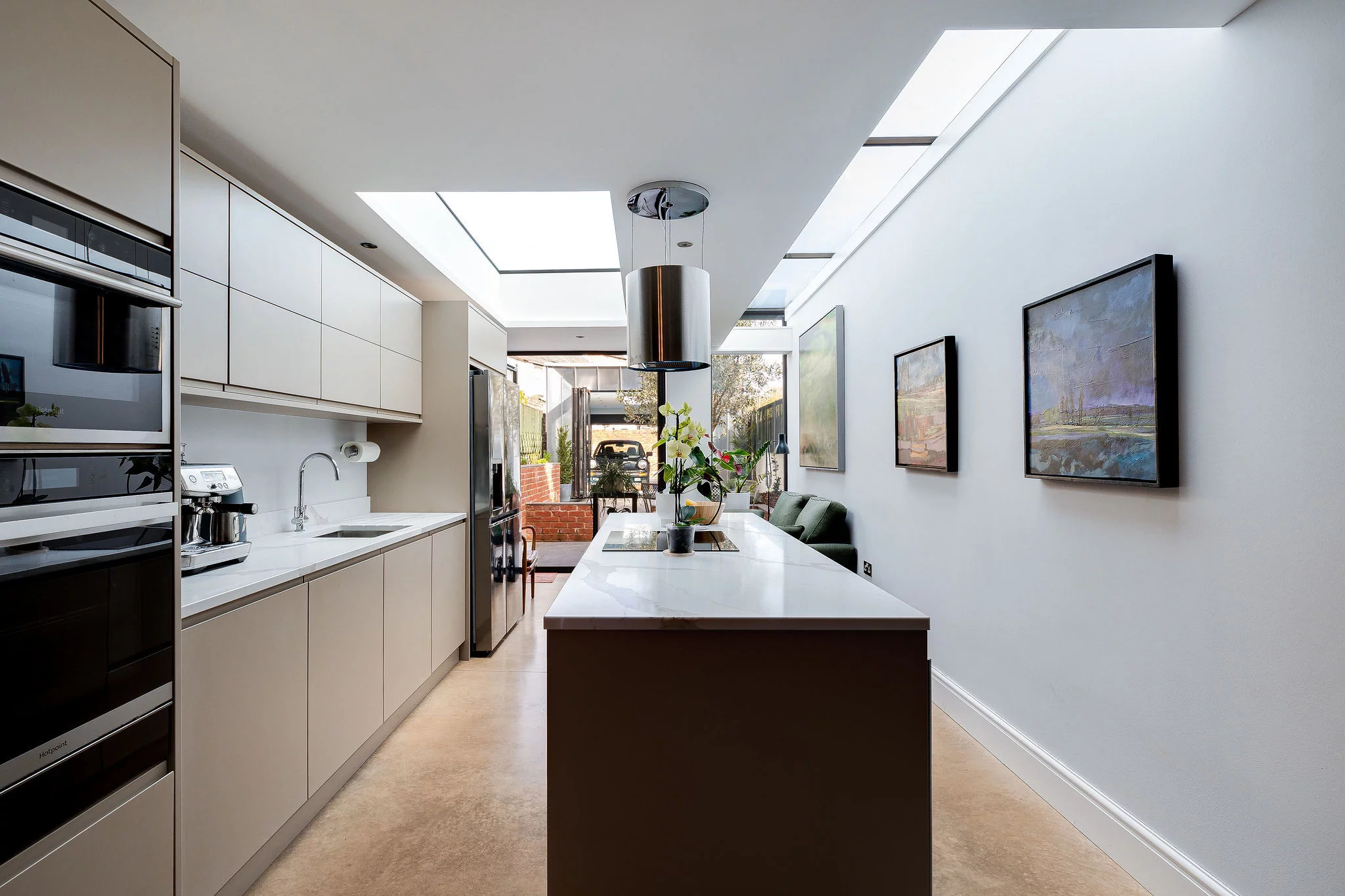 Narrow modern kitchen with long island, white countertops, skylights, stainless appliances and framed paintings on the right wall