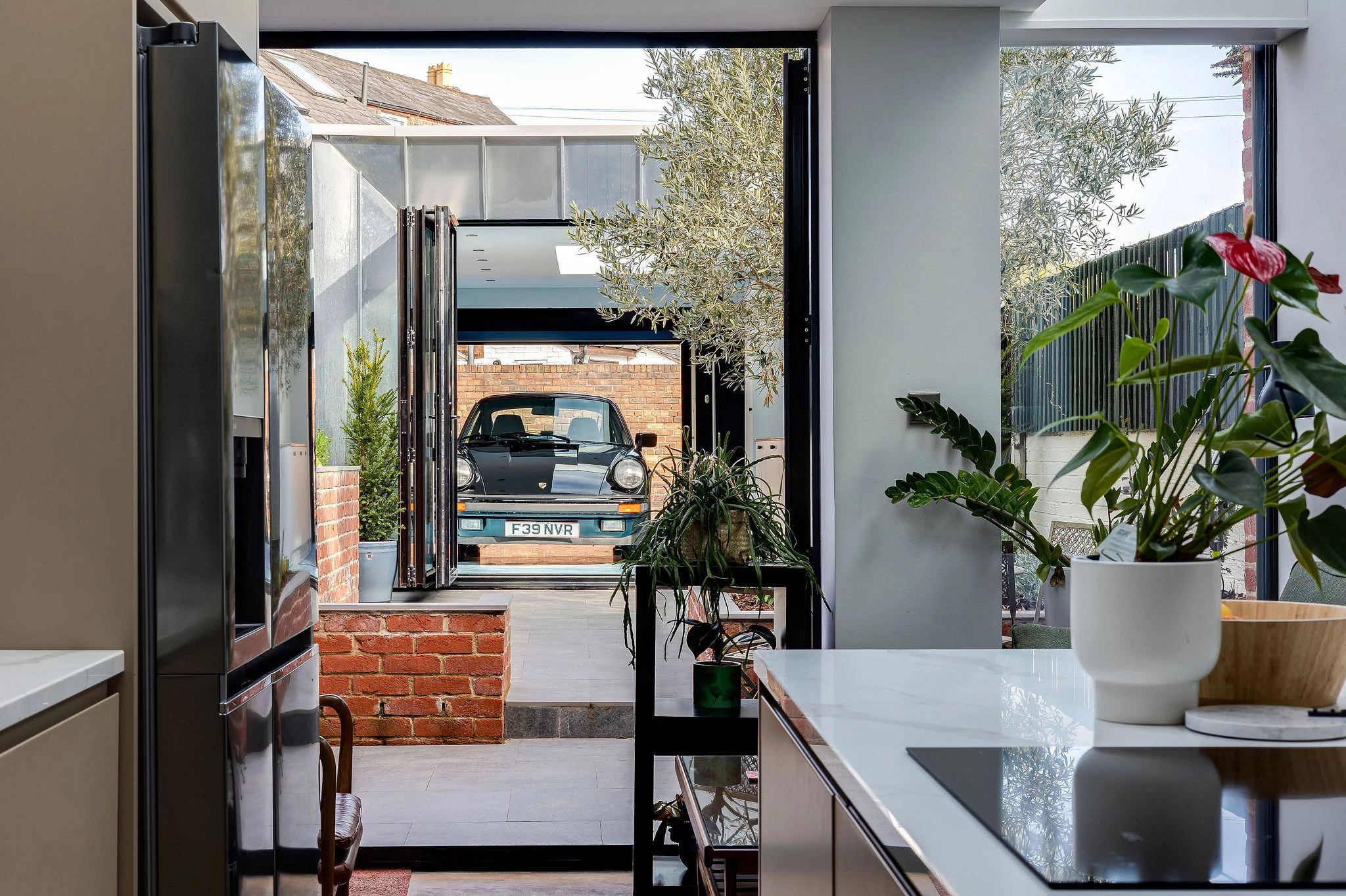 Modern kitchen interior looking through open glass doors to a garage with a vintage black Porsche and potted plants.