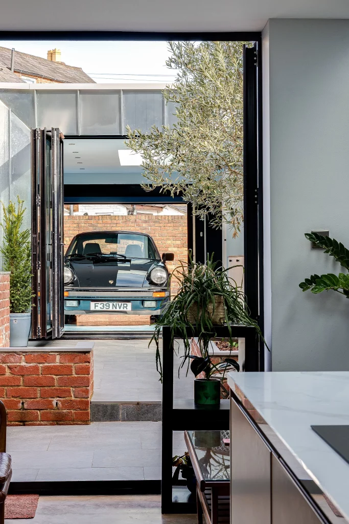 Classic black Porsche parked in a modern home extension, viewed through open folding glass doors with potted plants nearby.