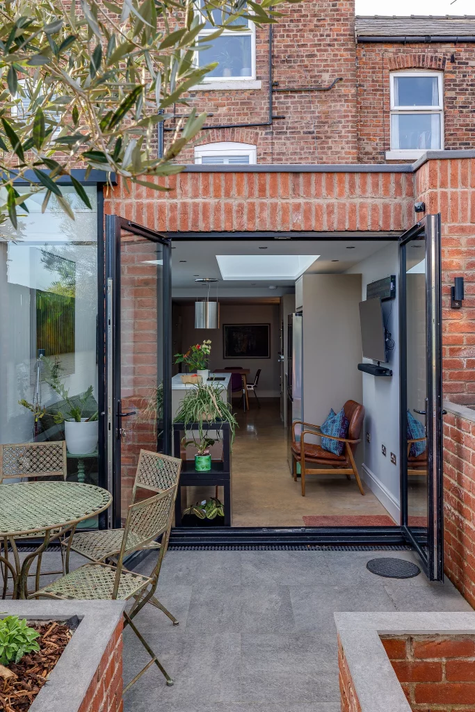 Open folding glass doors from a brick house onto a paved patio with metal bistro table and chairs, showing an interior with plants, a leather armchair and kitchen beyond.
