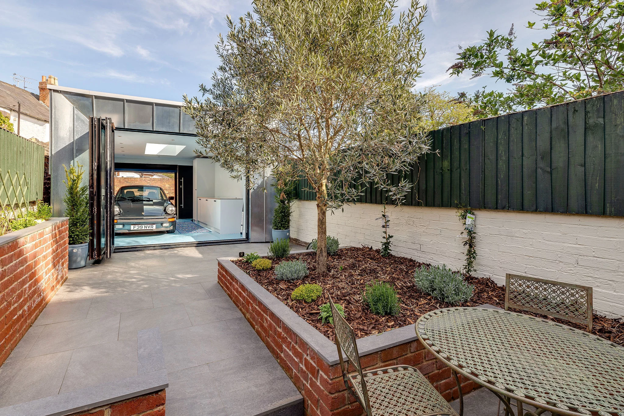 Rear garden with paved patio, raised brick planter and olive tree, metal bistro set, and a modern garage with a parked car.