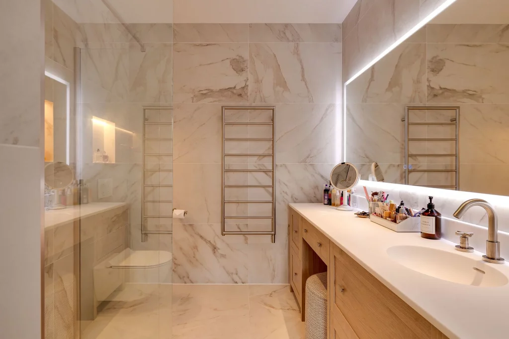 Modern marble bathroom with wall-mounted towel warmer, wood vanity, illuminated mirror, sink and toiletries on the countertop.