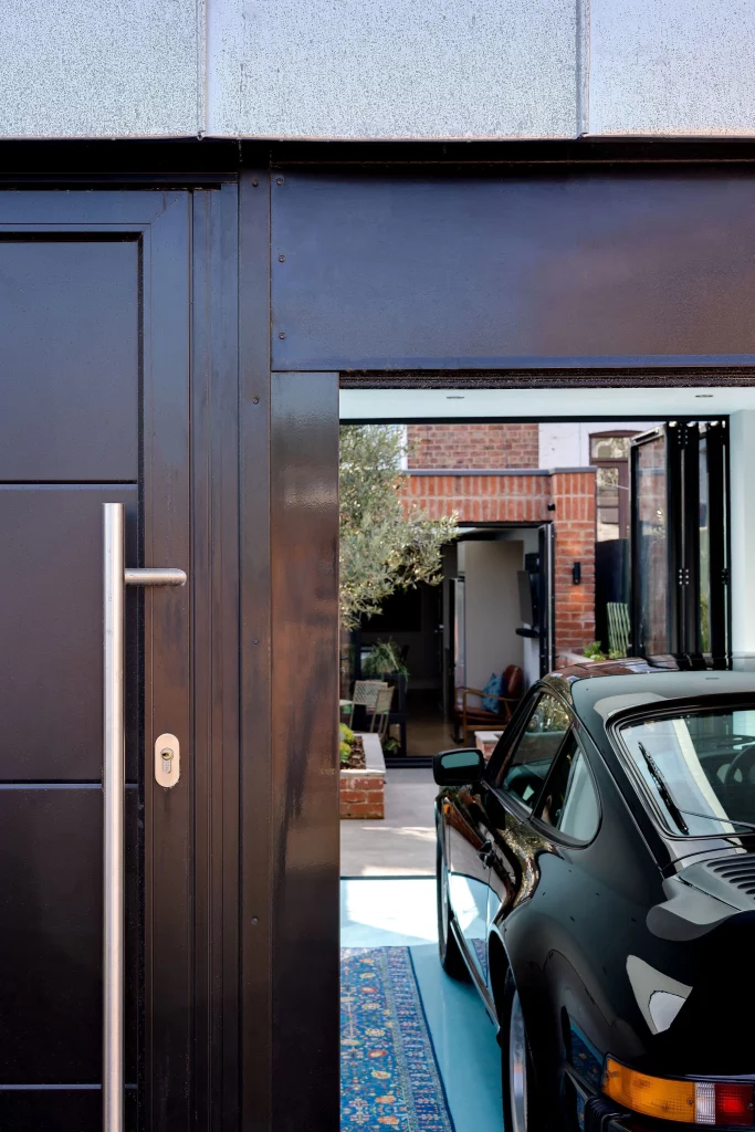 Black sports car parked inside a modern garage with open door revealing a brick-walled courtyard and indoor seating.