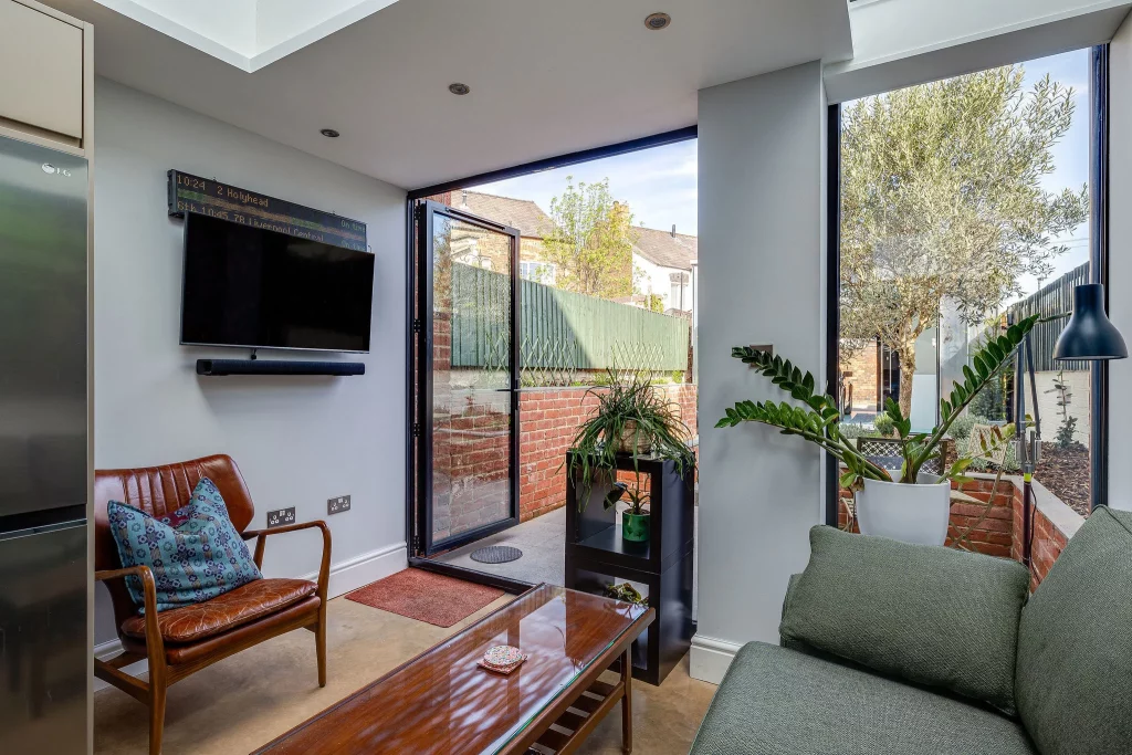 Sunlit living room with green sofa, leather armchair, wall-mounted TV, coffee table and open glass door to a small brick patio.
