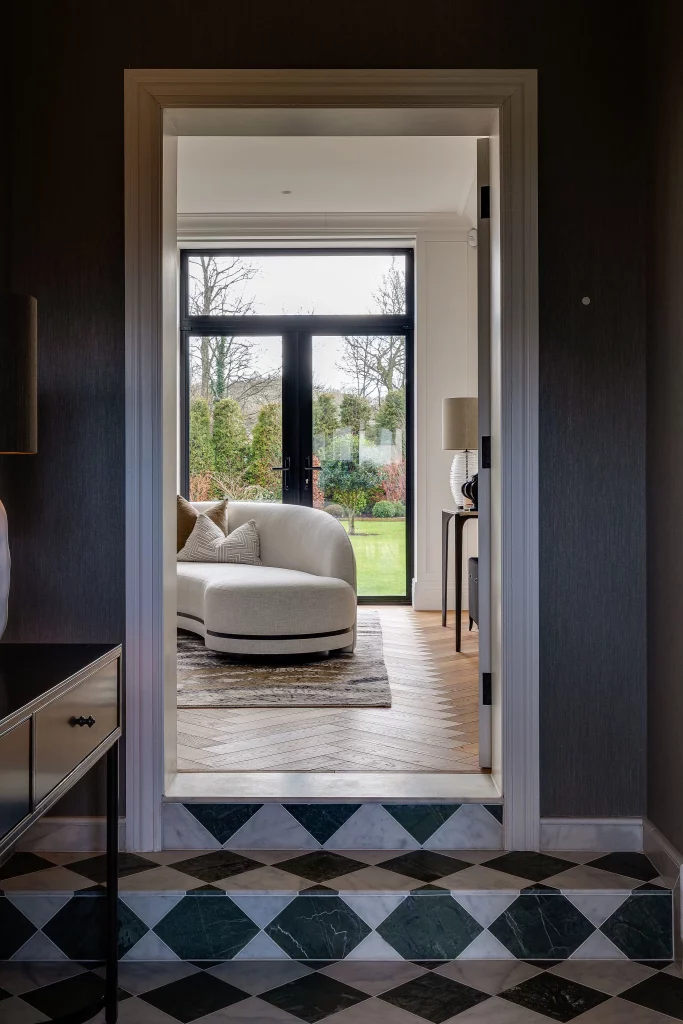 View through a doorway to a bright sitting room with a curved cream sofa, rug, parquet floor and glass doors opening to a garden.