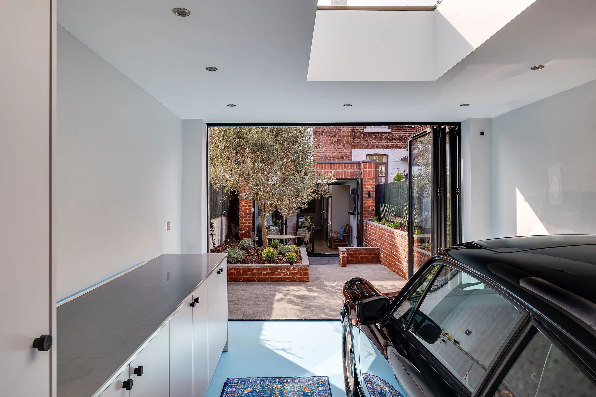 Modern garage with a black car, skylight, white cabinets and open folding doors leading to a small brick courtyard garden.