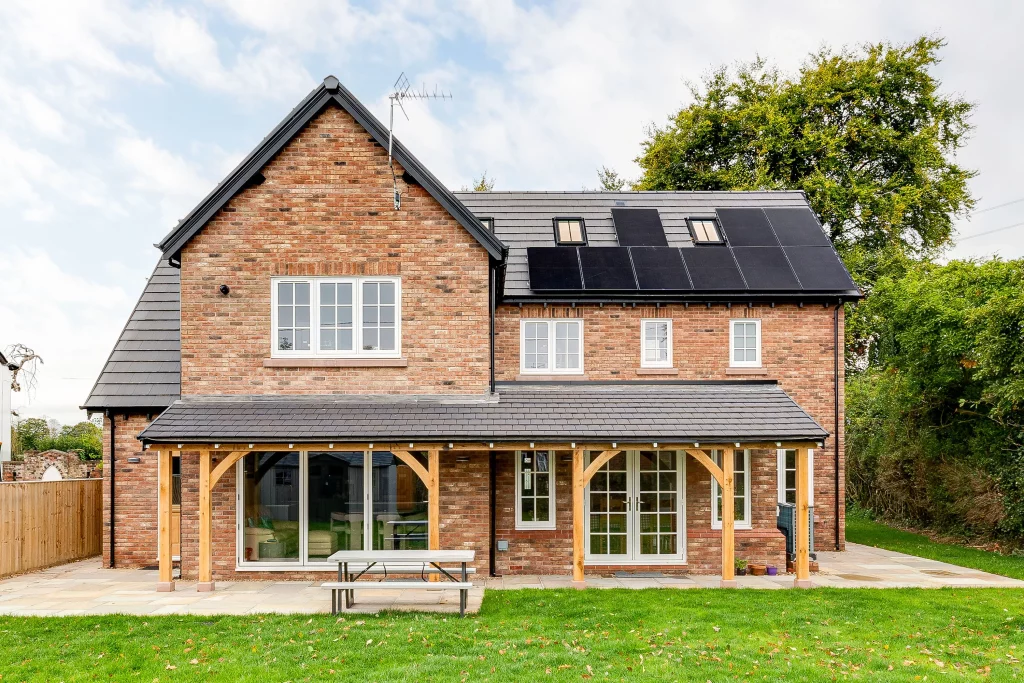 Two-storey redbrick house with slate roof, solar panels on the upper right roof, white-framed windows and a wooden porch over sliding patio doors.