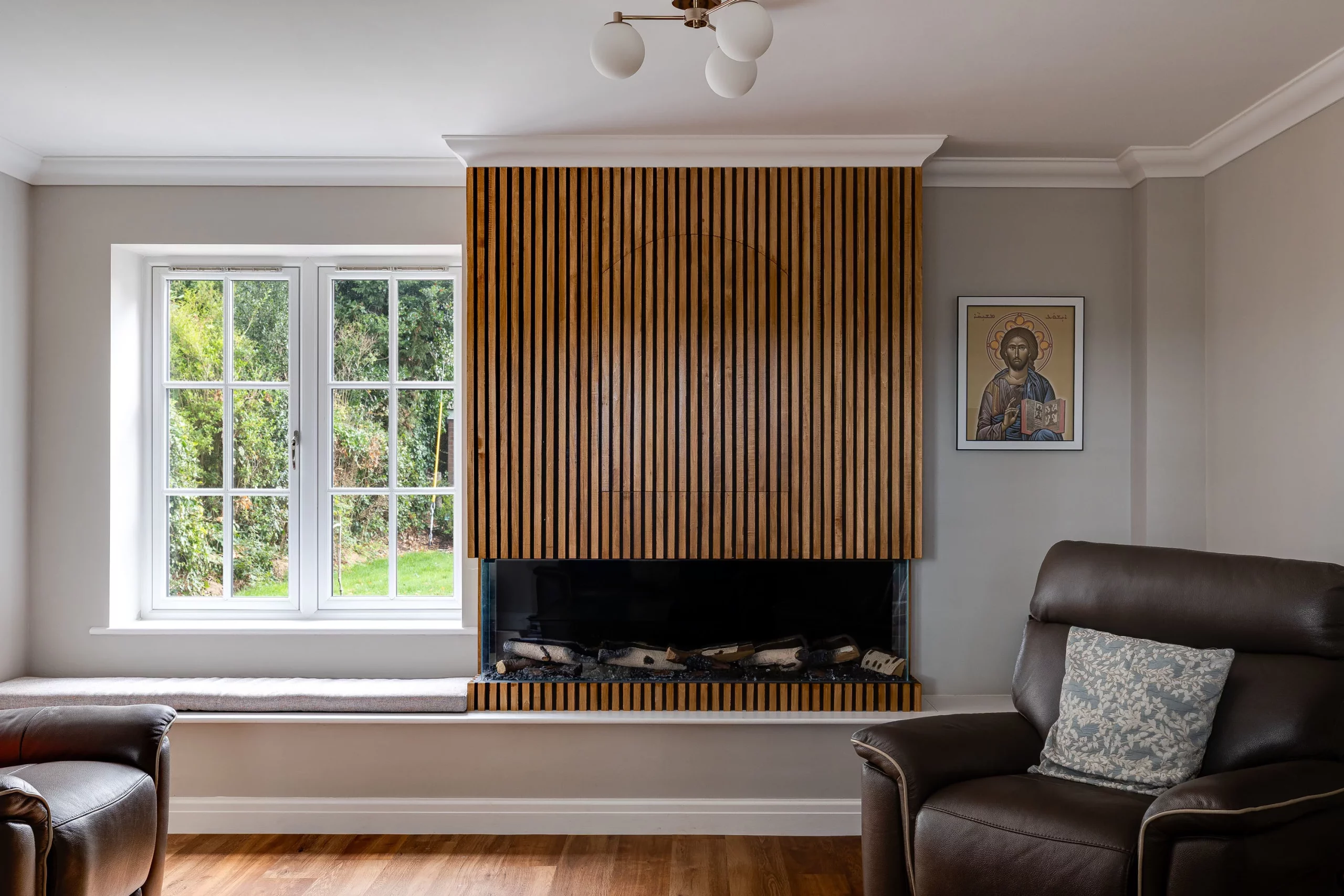 Living room with a vertical slat wooden fireplace surround between a white-framed window and a framed religious icon, leather armchairs at edges.