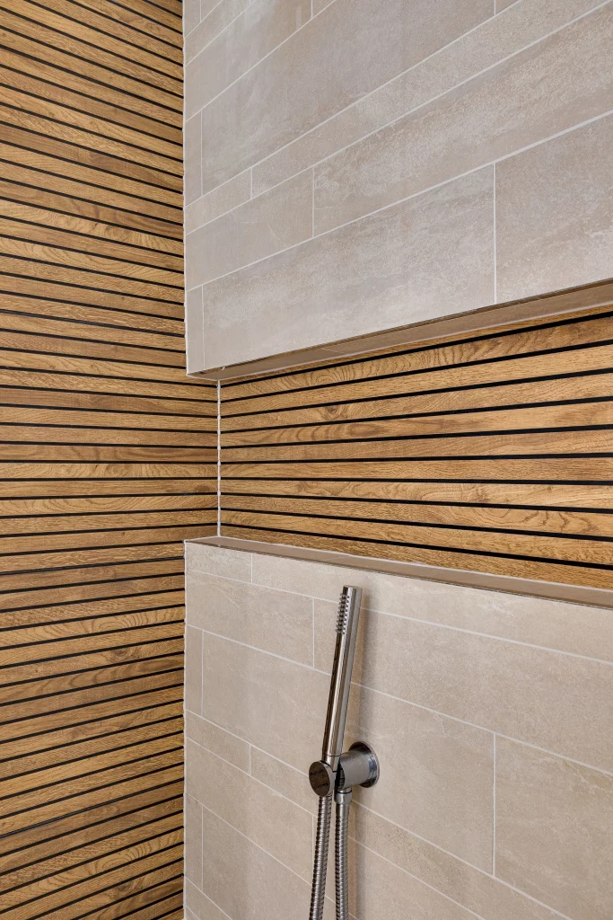 Shower wall with beige rectangular tiles and vertical chrome handheld showerhead against wood slat panelled corner walls