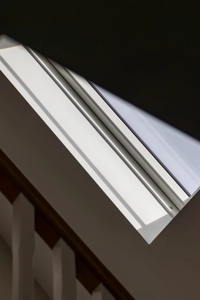 Slanted close-up of a white roof window frame and glass casting sharp diagonal shadows against a dark ceiling and stairwell