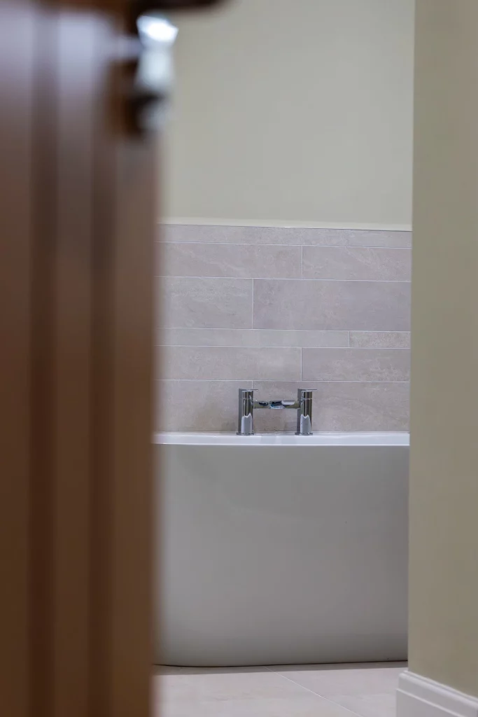Partially open door revealing a white bathtub with chrome faucet against beige tiled wall