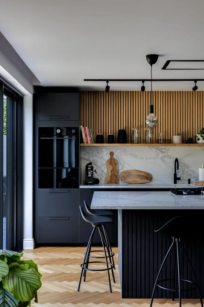 Modern kitchen with black cabinetry, marble island, wooden slatted backsplash, bar stools and pendant light over breakfast bar