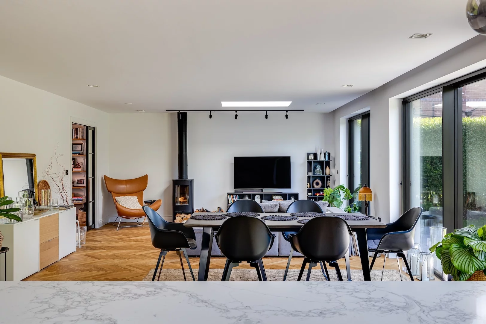 Open-plan dining and living area with a long black table and six black chairs, a wall-mounted TV, a leather lounge chair, wood-burning stove, and large sliding glass doors.
