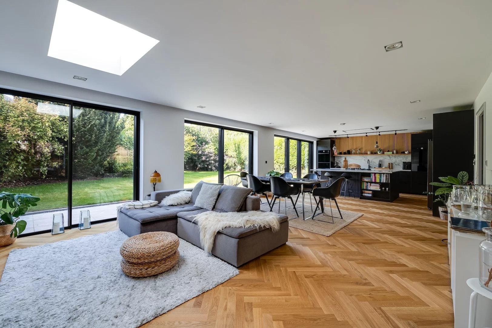 Bright open-plan living room with gray sectional, dining table, large sliding windows overlooking a green garden.