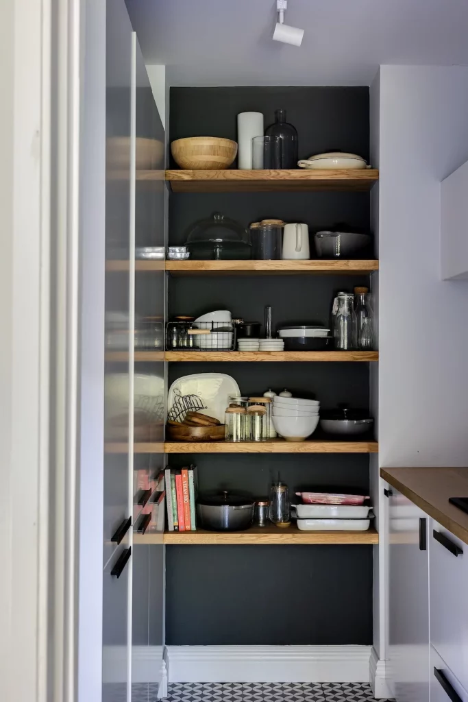Open kitchen shelving with wooden shelves against a dark back wall holding bowls, jars, dishes, cookware and cookbooks.