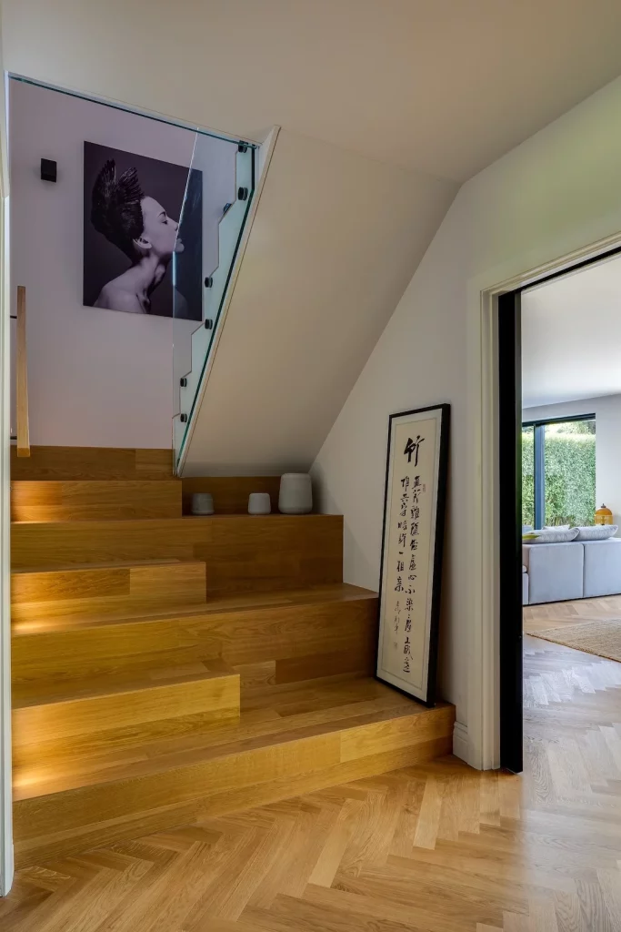 Oak staircase with glass balustrade, decorative vases, framed calligraphy leaning on wall and black-and-white portrait above.