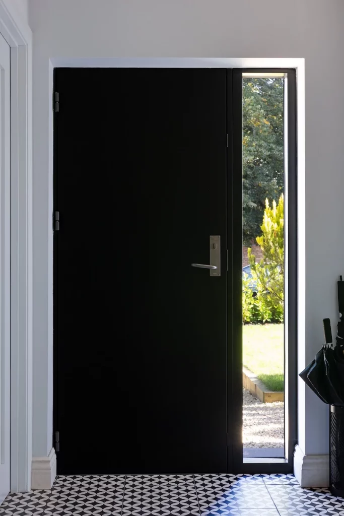 Tall matte black front door with narrow vertical sidelight showing sunlit garden, patterned black-and-white tiled floor, umbrella stand.