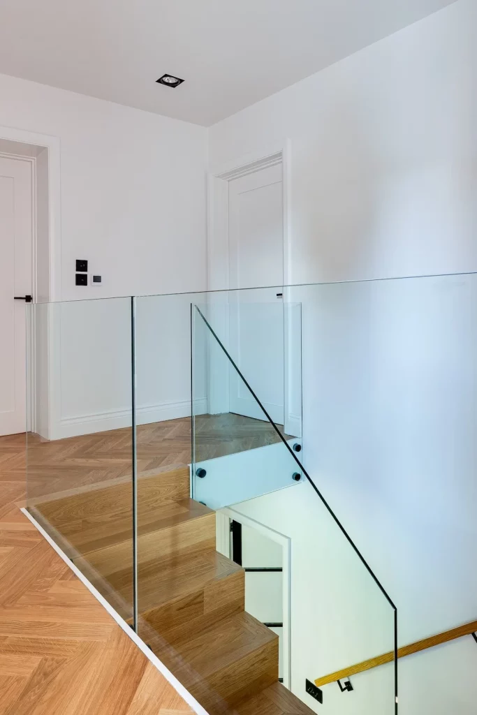 Minimal interior stairwell with oak steps, glass balustrade and white walls leading to a closed white door on the landing
